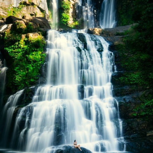 A mesmerizing view of Nauyaca Waterfalls in Costa Rica, with cascading tiers of water flowing into a pristine pool below. A lone woman sits on a rock at the base of the waterfall, enjoying the serene beauty and refreshing mist. This natural gem, surrounded by lush green foliage, is one of the top things to do in Uvita, offering visitors a chance to connect with nature and experience the tranquility of Costa Rica’s wilderness.