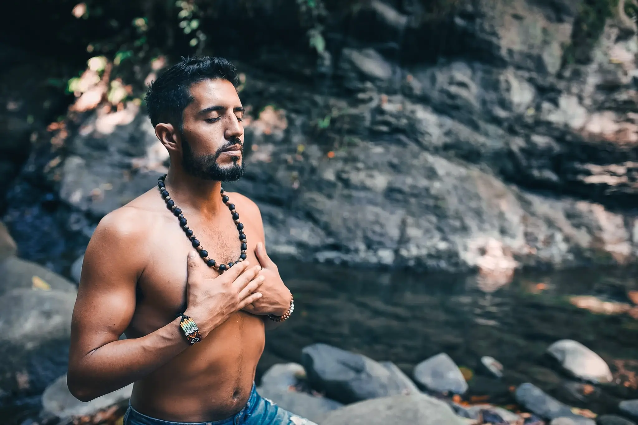A man meditates with his hands on his chest, standing by a tranquil river and rocky landscape at Vayu Retreat Villas. This serene moment captures the essence of relaxation and mindfulness in one of the best hotels in Uvita, Costa Rica, offering a perfect setting for a peaceful jungle retreat.