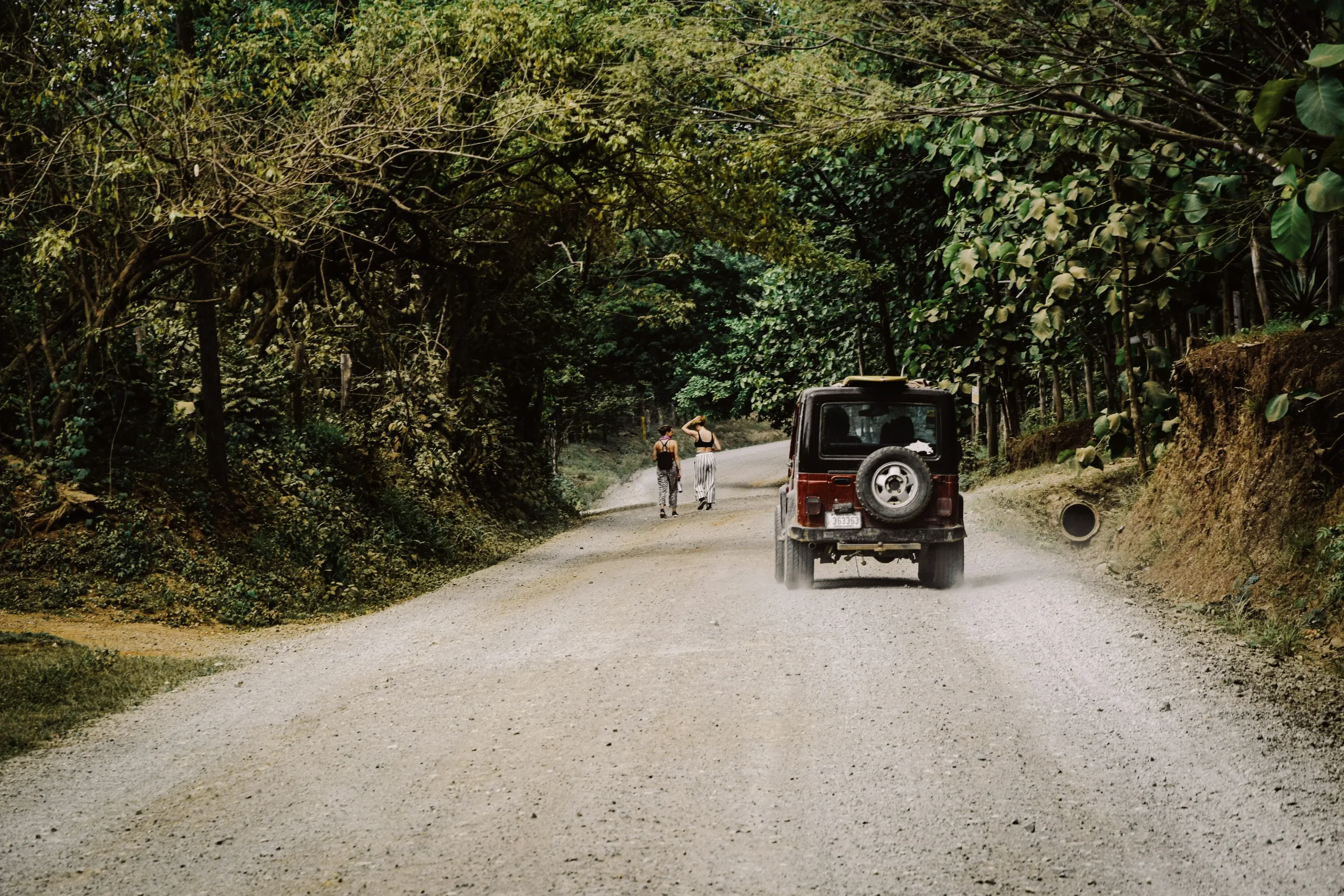 Rental car driving on a dirt road surrounded by lush greenery near a boutique hotel in Uvita, Costa Rica, with two people walking ahead. Ideal for a wellness retreat in Uvita, Costa Rica.