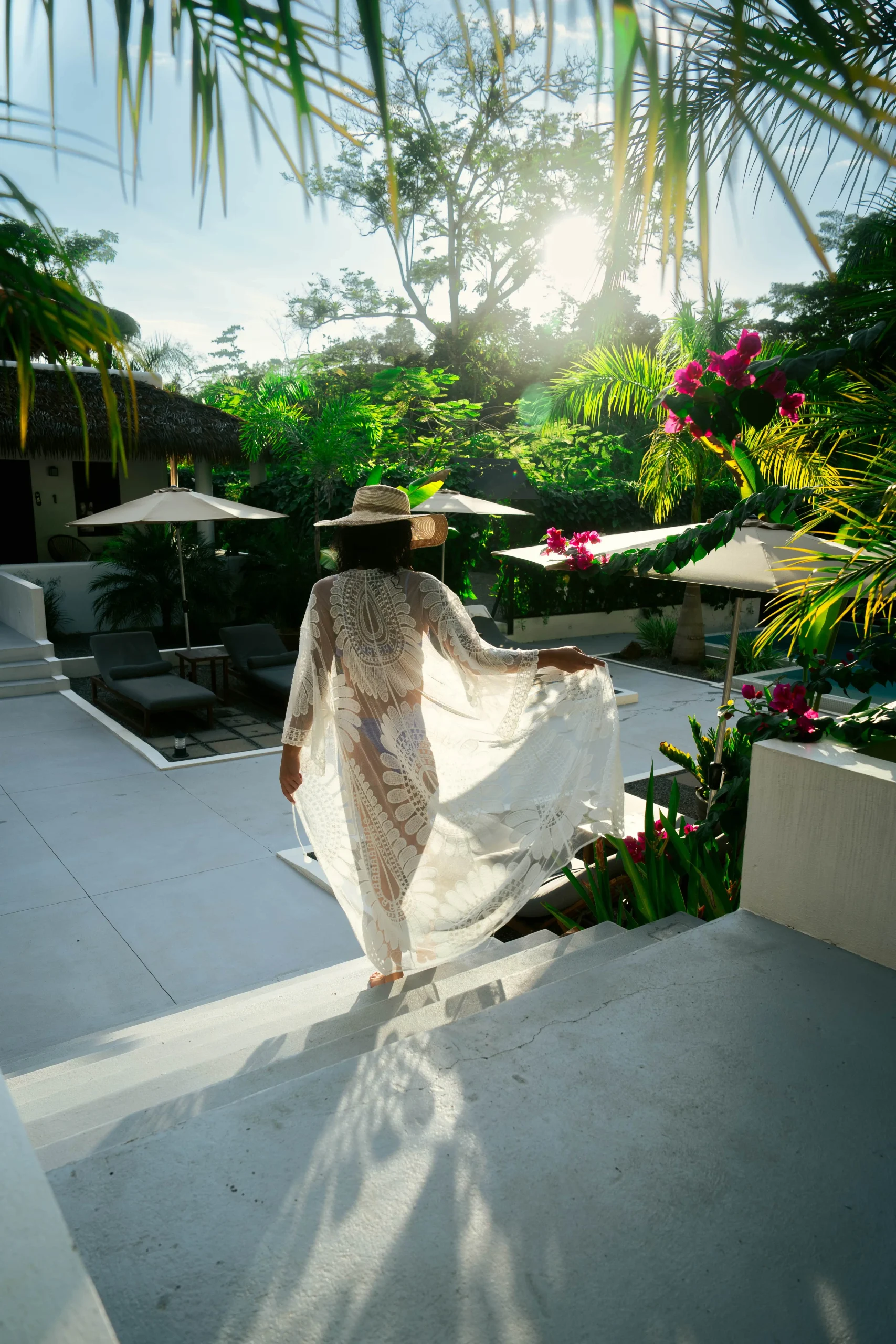 A woman in a flowing white cover-up and sun hat walks down the steps at Vayu Retreat Villas on a sunny day, surrounded by vibrant greenery and tropical flowers. This idyllic scene captures the essence of one of the best hotels in Uvita, Costa Rica, offering a perfect blend of luxury and natural beauty in a jungle retreat setting.