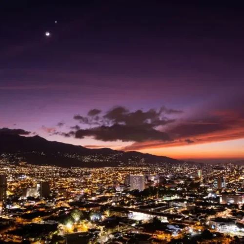 Visiting San José, Costa Rica: A breathtaking aerial view of the city's skyline at dusk, with vibrant city lights and a stunning sunset over the mountains.