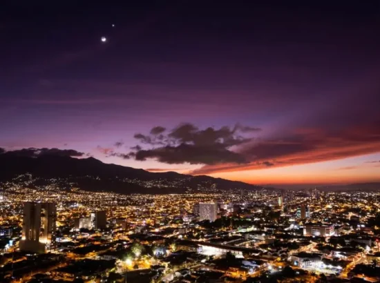 Visiting San José, Costa Rica: A breathtaking aerial view of the city's skyline at dusk, with vibrant city lights and a stunning sunset over the mountains.