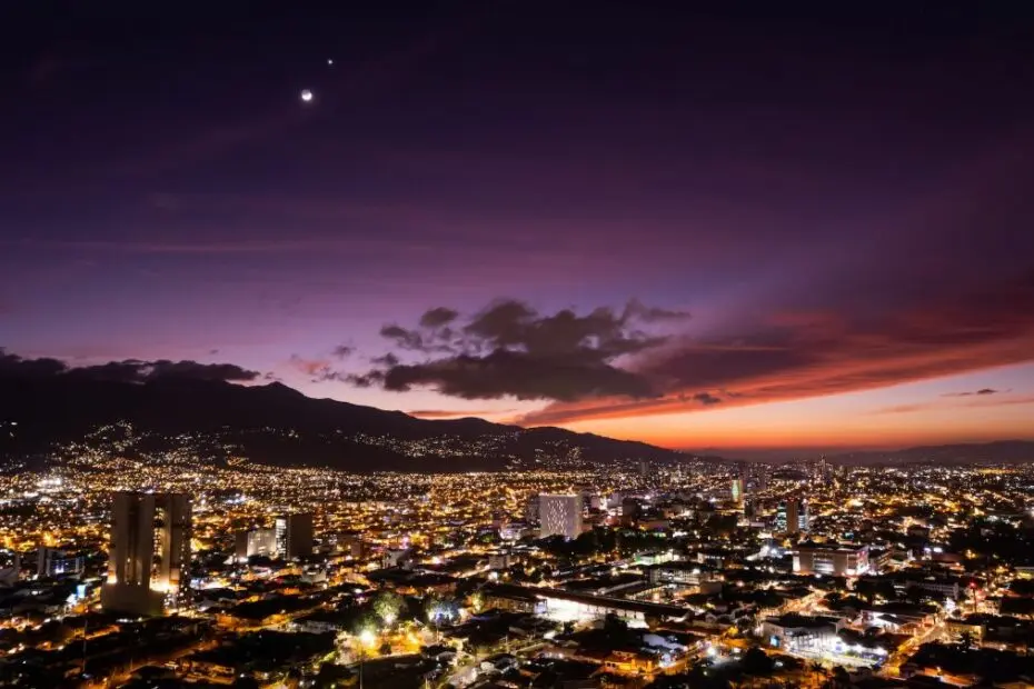 Visiting San José, Costa Rica: A breathtaking aerial view of the city's skyline at dusk, with vibrant city lights and a stunning sunset over the mountains.