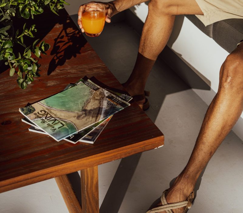 A person sitting on a patio at Vayu Retreat Villas, the best resort in Costa Rica, holding a glass of orange juice, with travel magazines on a wooden table and lush green plants in the background.